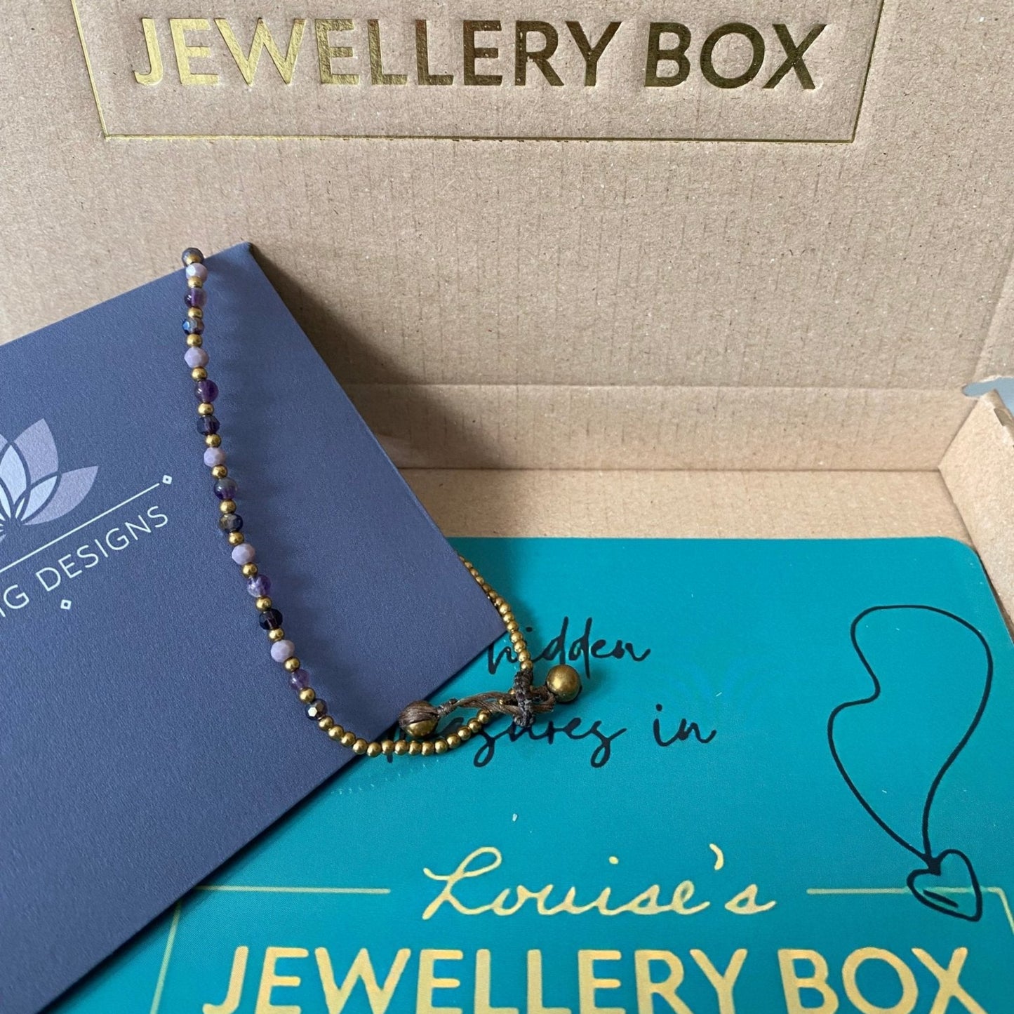 Amethyst crystal healing anklet for anxiety with gold beads, over a branded postal box.