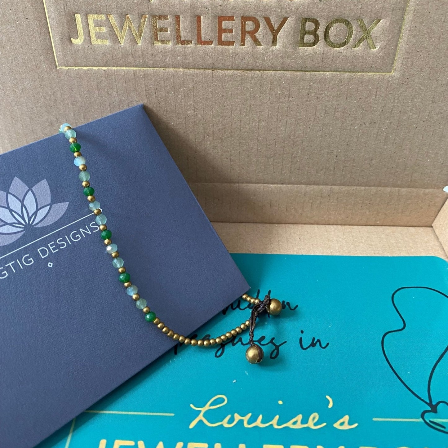 Green and gold aventurine anklet for energy by Rigtig Designs resting on packaging.
