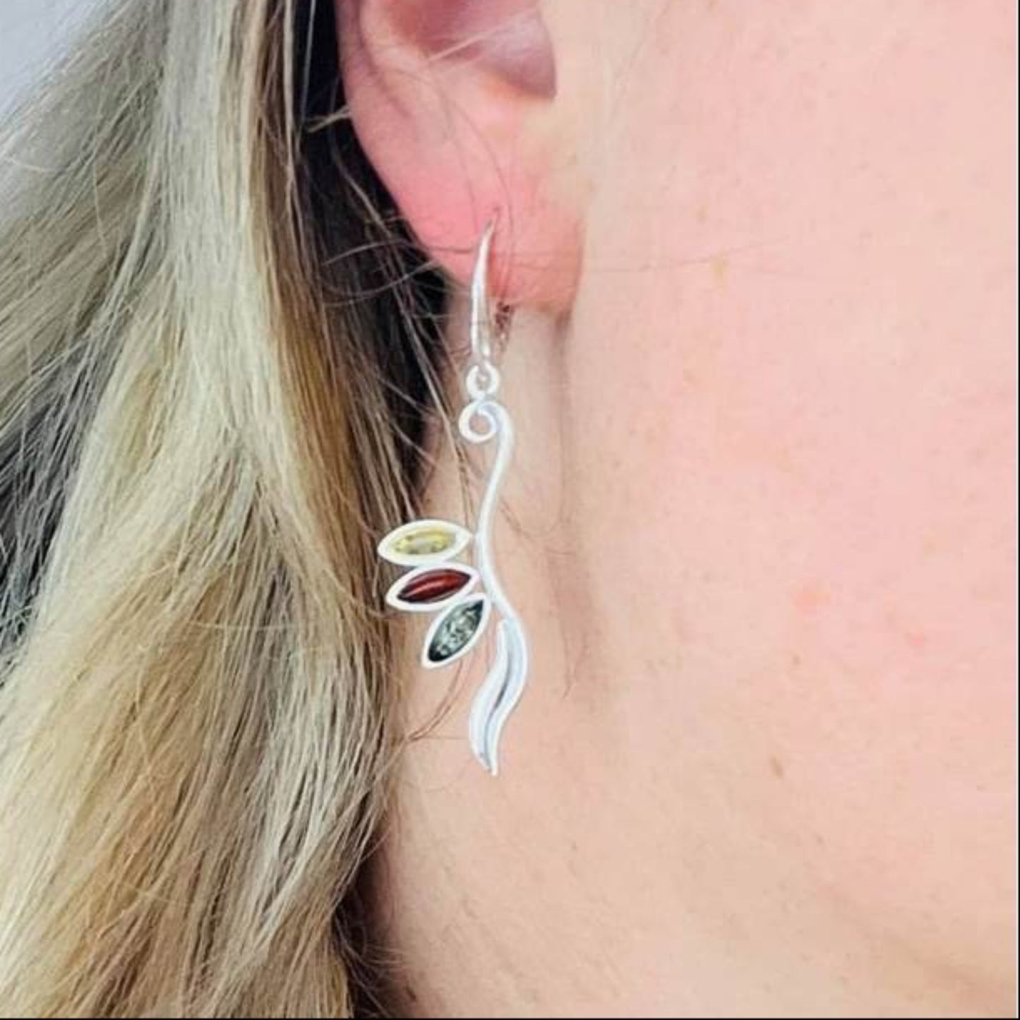 Sterling silver earrings with mixed Baltic amber by are styled on a person's ear.