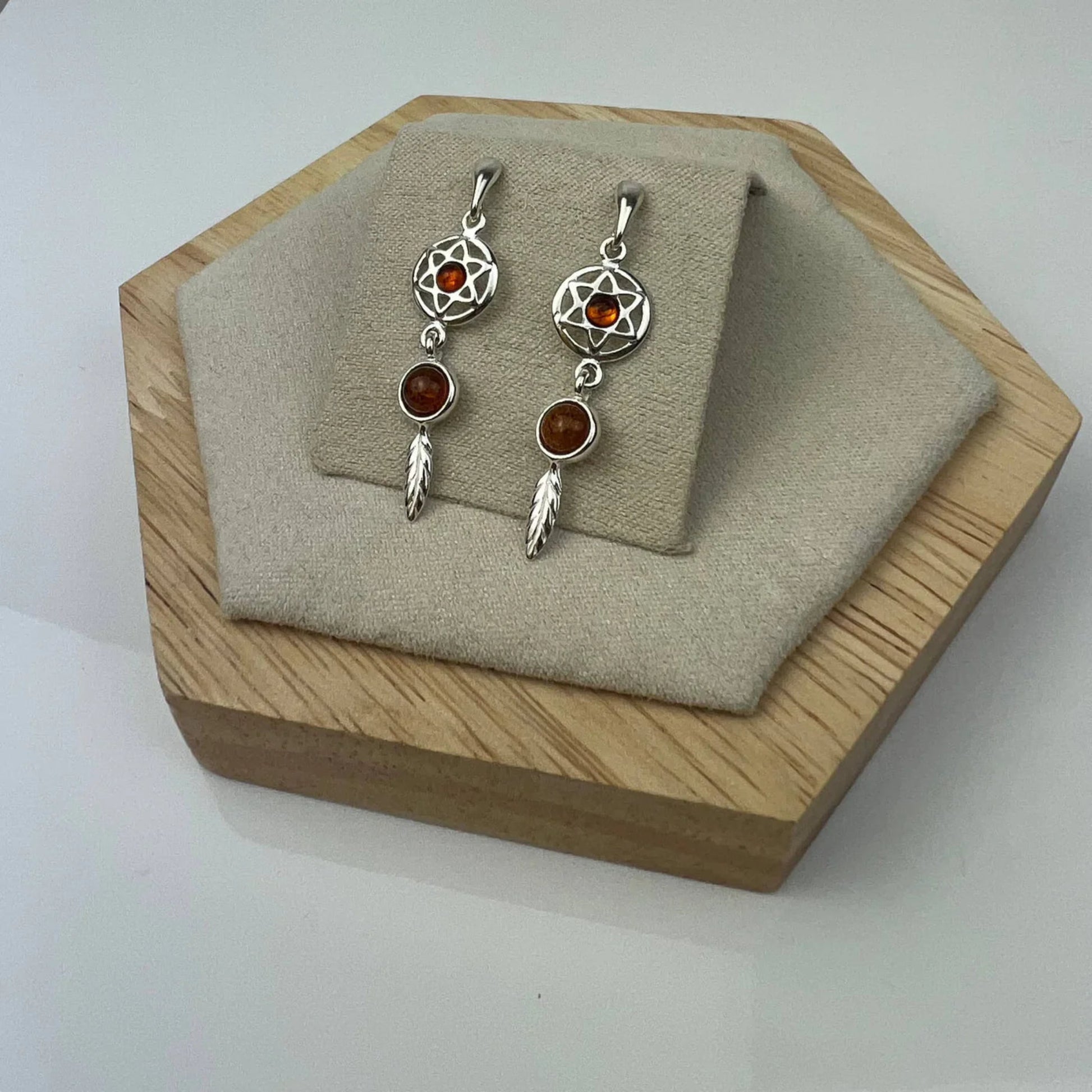 Sterling silver dreamcatcher earrings with cognac Baltic amber by Glow Silver, displayed on a layered wooden and fabric stand.